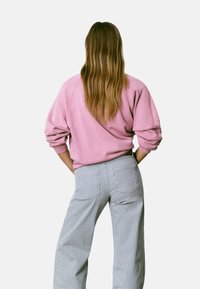 Persona con cabello largo castaño claro, vistiendo un sudadera rosa y pantalones a rayas gris claro, de pie con las manos en las caderas, mirando hacia el frente.