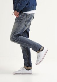 Farkut, joissa haalistunut sininen pesu, kapeahko istuvuus ja näkyvät ompelusaumat, yhdistettynä valkoisiin tennareihin, joissa on punaiset korosteraitoja.