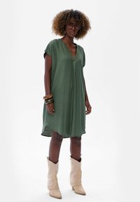 Oliwkowa sukienka z dekoltem w serek i krótkimi rękawami. Wykonana z miękkiego materiału, charakteryzująca się luźnym fasonem i zaokrąglonym dołem. Zestawiona z beżowymi kowbojkami.