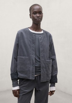Staande persoon met een gelaagde donkergrijze casual jas, bijpassende broek en een overhemd met lange mouwen en witte manchetten tegen een effen lichte achtergrond.