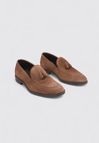 Mocasines de ante en marrón con punta afilada y borlas decorativas. Los zapatos cuentan con recortes laterales y una suela plana.