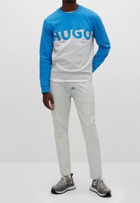 Hellblauer und weißer Sweatshirt mit auffälligem "HUGO"-Schriftzug, kombiniert mit hellen Jeans und grauen Sneakers mit schwarzen Akzenten.