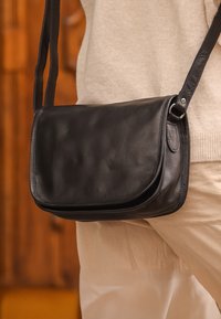 Borsa a tracolla nera in pelle con una superficie liscia, chiusura a ribalta e tracolla sottile e regolabile. Presenta hardware minimal e un design elegante.