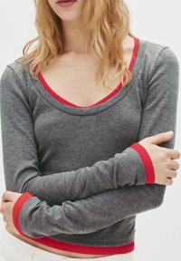 Tricou lung gri cu mâneci lungi și decolteu rotund. Accente roșii pe guler și manșete. Fabricat dintr-un material moale, având un design ajustat.