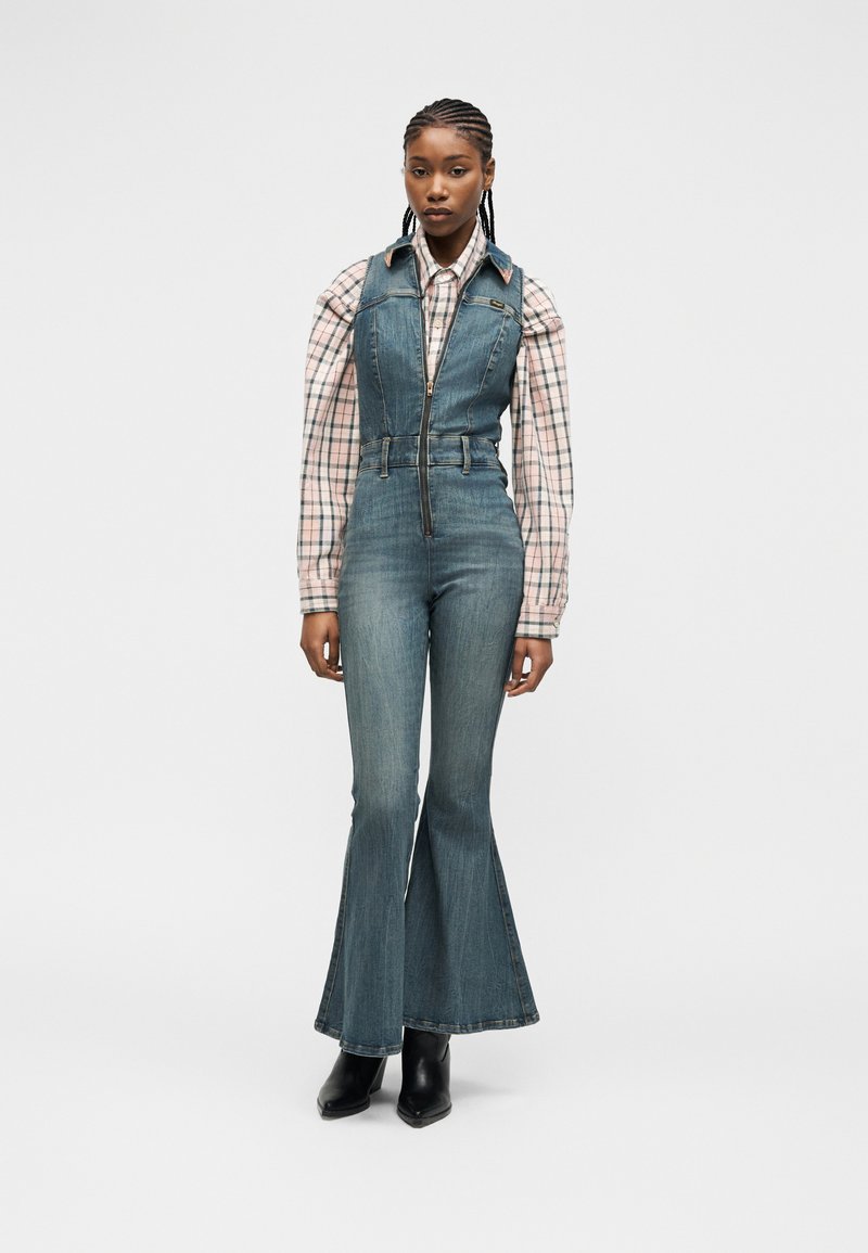 Γυναίκα με πλεγμένα μαλλιά που φοράει ολόσωμη φόρμα τζιν με φερμουάρ και καμπάνα στο κάτω μέρος, από μέσα ροζ πουκάμισο με καρό και μαύρα μποτάκια μέχρι τον αστράγαλο, στέκεται μπροστά σε λευκό φόντο.