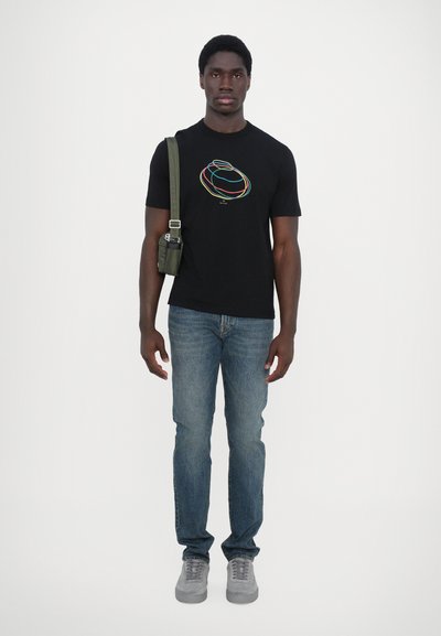 T-shirt noire avec un design abstrait coloré, associée à un jean bleu clair et des baskets grises. Le modèle porte un sac bandoulière vert foncé.