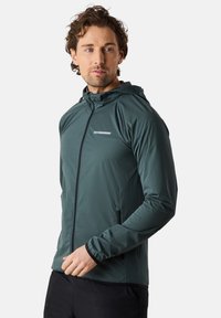 Giacca con cappuccio di colore verde scuro, realizzata in tessuto leggero. Caratterizzata da una zip frontale, tasche laterali e un'accenno riflettente sul petto.