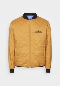Veste bomber matelassée jaune avec un col et des poignets en rib de couleur noire. Elle possède une fermeture éclair à l'avant et un logo "Obey" brodé sur la poitrine.
