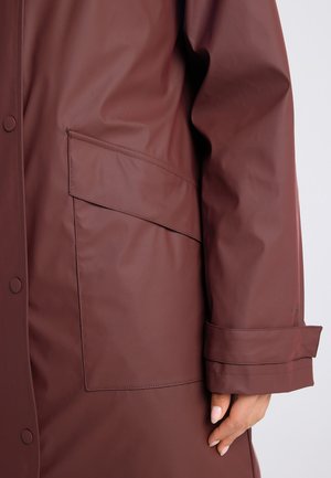 Brauner wasserdichter Mantelärmel und Tasche mit Klappe, geknöpfter Ärmelbund, getragen am Arm mit sichtbarer Hand vor einfarbigem Hintergrund.