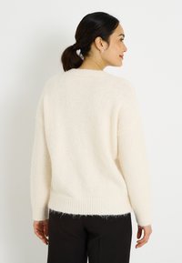 Suéter de punto color crema con textura suave, hombros caídos y dobladillo acanalado. Cuenta con mangas largas y holgadas y un ajuste relajado.