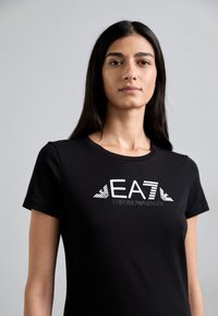 Svart kortermet t-skjorte laget av bomull, med en fremtredende hvit logo med geometriske detaljer "EA7" og "EMPORIO ARMANI" på forsiden.