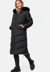 Giacca puffa nera con un design lungo e trapuntato, dotata di cappuccio foderato in pelliccia, chiusura con zip e tasche laterali. Indossata con stivali neri con lacci.