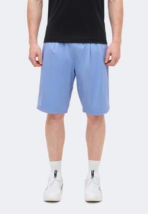 Άνδρας που φοράει ανοιχτό μπλε σορτς μέχρι το γόνατο, λευκά αθλητικά παπούτσια, λευκές κάλτσες και μαύρο πουκάμισο, στέκεται μπροστά από απλό φόντο.