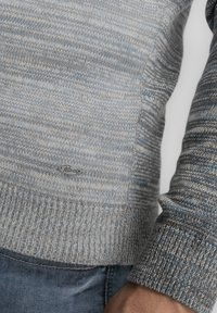 Strickpullover in hellgrauen und blauen Farbtönen, mit einem strukturierten Muster und einem gerippten Saum. Kleines Logotag in der Nähe des Saums sichtbar.