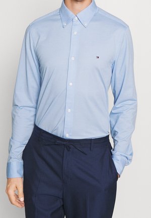 Mężczyzna noszący jasnoniebieską koszulę na guziki z małym logo na piersi oraz ciemnogranatowe spodnie ze sznurkiem w pasie.