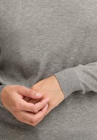 Gray sweater made of soft fabric, featuring a crew neckline, ribbed cuffs, and hem. Hands clasped gently in front of the body.