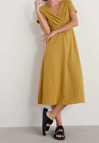 Musztardowa sukienka w białe kropki, z krótkimi rękawami i dekoltem w serek. Połączona z czarnymi sandałami z podwójnym zapięciem.