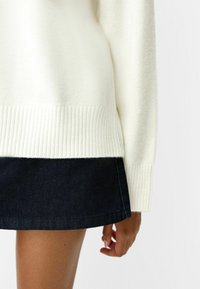 Sweter w kolorze kremowym, wykonany na drutach, z prążkowanym dołem i mankietami, zestawiony z ciemną spódnicą dżinsową. Materiał wydaje się miękki i teksturowany.