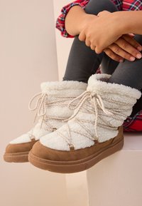 Bottes montantes blanches doublées de fourrure, avec des panneaux en suède beige et des lacets. Bout arrondi et semelle en caoutchouc. Tige texturée offrant chaleur et confort.