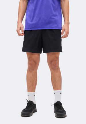 Férfi fekete sport rövidnadrágban, fehér Nike zokniban, fekete sportcipőben és lila pólóban, egyszínű világos háttér előtt áll.