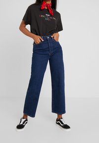 Černé tričko s krátkým rukávem a červeným a zeleným potiskem "Levi Strauss & Co.", doplněné o tmavě modré široké džíny s vysokým pasem a černé tenisky.
