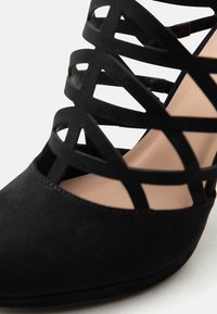 Μαύρο παπούτσι με ψηλό τακούνι, κλειστή μυτερή μύτη και πολλούς λεπτούς ιμάντες που σχηματίζουν γεωμετρικό μοτίβο πάνω στο πόδι.