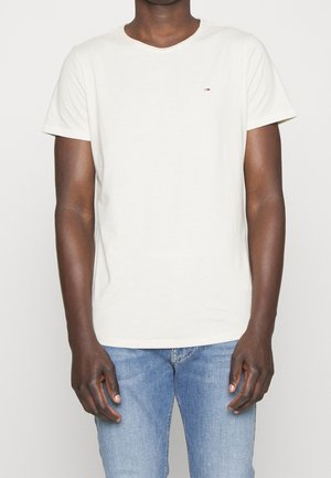 Mężczyzna ubrany w prostą białą koszulkę z krótkim rękawem i jasnoniebieskie dżinsy, stojący na tle jednolitego jasnego tła.