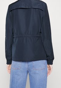 Mörkblå lättviktsjacka med insvängd midja och elastiska mudd, med bakre ok och slät textur, bärs över ljusblå jeans.