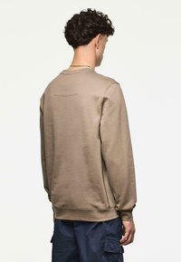 Bruine sweatshirt met ronde halslijn, ribbelmanchetten en een detail van een achternaad. Draagt met donkere cargobroek. Glad, katoenachtig textuur.