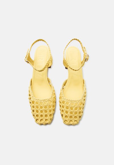 Κίτρινα πλεκτά σανδάλια με λουράκι στο πίσω μέρος, τετράγωνες μύτες και ρυθμιζόμενα λουράκια στον αστράγαλο, παρουσιάζονται σε λευκό φόντο.