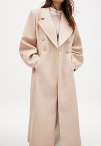Dvojradový, dlhý béžový kabát s širokým klopom, oversized rukávmi a textúrovanou tkaninou. Obsahuje predné gombíky a bočné vrecká.