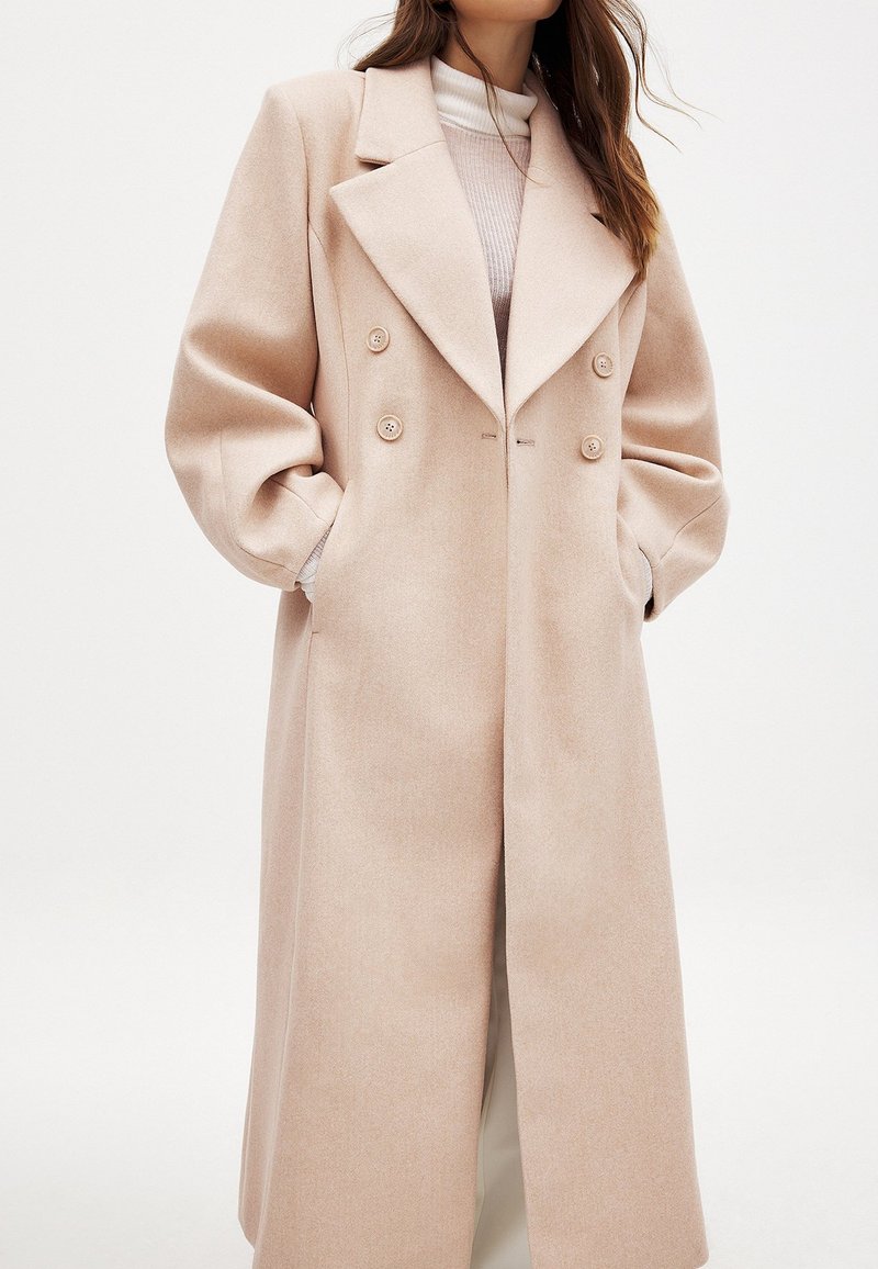 Dvojradový, dlhý béžový kabát s širokým klopom, oversized rukávmi a textúrovanou tkaninou. Obsahuje predné gombíky a bočné vrecká.