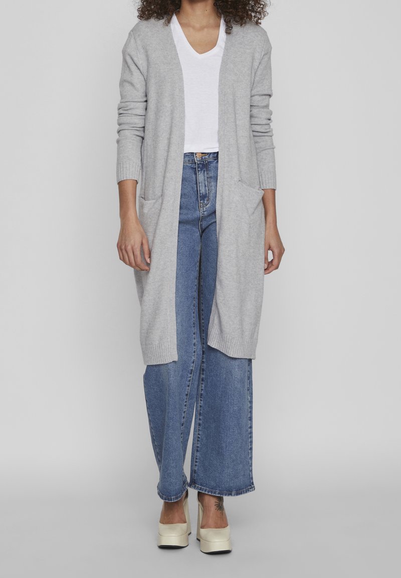 Ljusgrå stickad cardigan med långa ärmar och sidofickor, bärs över en vit V-ringad skjorta och matchas med vida blå jeans.