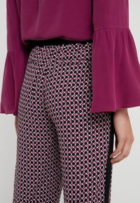Paarse blouse met uitlopende mouwen, gecombineerd met geprinte broek met roze, witte en zwarte geometrische vormen en zijaccenten.