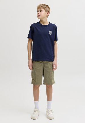 Ragazzo con maglietta blu navy con piccolo logo bianco, pantaloncini cargo kaki, calzini bianchi e scarpe da ginnastica bianche, guarda a sinistra.