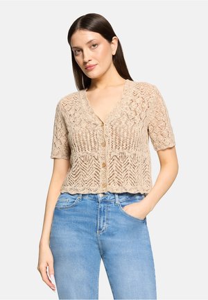 Jeune femme aux cheveux bruns lisses portant un haut en maille beige à manches courtes boutonné et un jean taille haute bleu clair, main dans la poche.