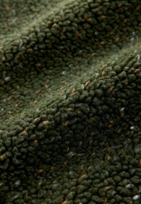 Primo piano di tessuto in pile verde con una superficie testurizzata e soffice e piccole fibre marroni e bianche distribuite qua e là.