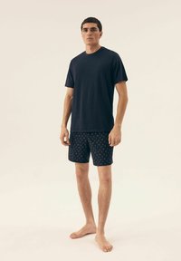 Granatowa koszulka z miękkiego materiału, w połączeniu z granatowymi szortami w wzory z żółtymi akcentami. Model stoi boso na neutralnym tle.