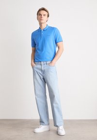 Polo de culoare albastră, fabricat din bumbac, asociat cu blugi deschiși la culoare și adidași albi. Polo-ul are guler și mâneci scurte.