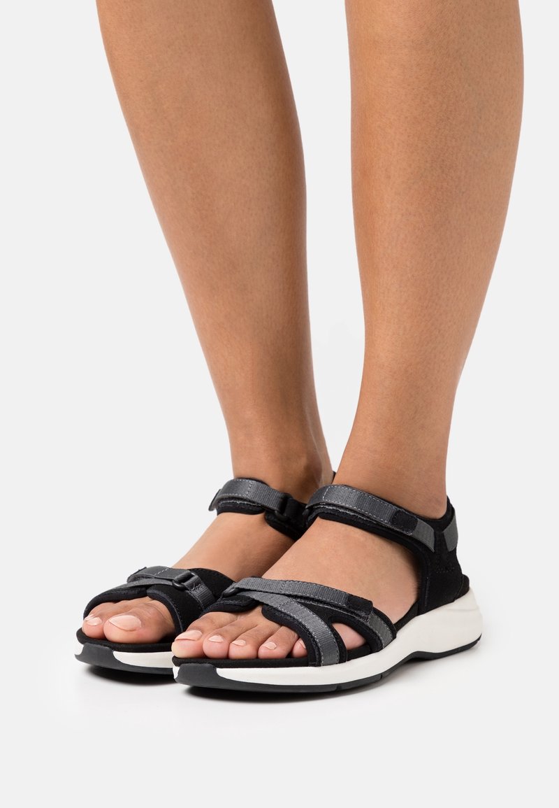 Jambes nues portant des sandales noires et grises à bouts ouverts avec des brides réglables et des semelles blanches épaisses sur un fond clair.