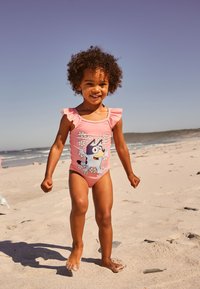 Rožinės spalvos vientisas maudymosi kostiumėlis su raukinukais ant pečių, animacinio personažo dizainas ir gėlių motyvai; vaikas žaidžia ant smėlėtos paplūdimio dalies.