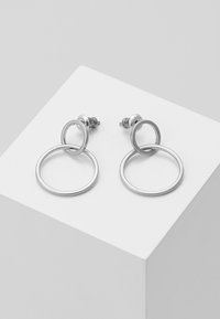 Boucles d'oreilles en argent avec des designs circulaires entrelacés. Texture métallique lisse, dotées d'un fermoir à tige pour un port sécurisé. Présentées sur un piédestal blanc.