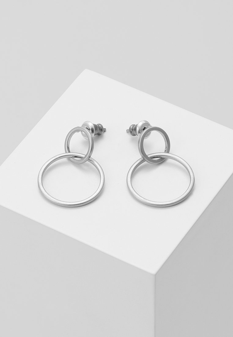 Boucles d'oreilles en argent avec des designs circulaires entrelacés. Texture métallique lisse, dotées d'un fermoir à tige pour un port sécurisé. Présentées sur un piédestal blanc.