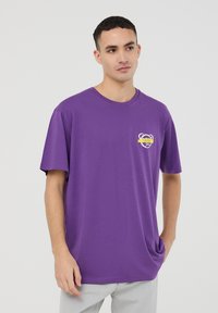 T-shirt viola a maniche corte in cotone, con scollatura rotonda e una piccola grafica di un orsetto con accenti gialli sul lato sinistro del petto.
