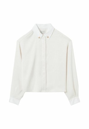 Donna con camicetta bianca abbottonata e pantaloni beige a gamba larga che tiene una borsa a tracolla in pelle marrone scuro con coulisse, in piedi su uno sfondo neutro.