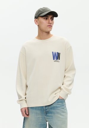 Młody mężczyzna ubrany w beżową koszulę z długim rękawem z logo "WW" i napisem, jasnoniebieskie dżinsy oraz ciemnozieloną czapkę z daszkiem, patrzący w bok.