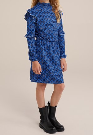 Kind trägt ein langärmeliges blau gemustertes Kleid mit Rüschen und schwarze Knöchelstiefel und steht vor einem einfarbigen beigen Hintergrund.