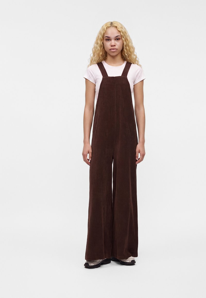 Jeune femme aux longs cheveux blonds bouclés portant un t-shirt blanc et une salopette large en velours côtelé marron, debout devant un fond uni.