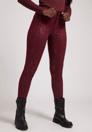Kvinde iført skinnende bordeaux teksturerede leggings kombineret med sorte støvler til midt på læggen, stående med hænderne, der justerer taljebåndet.