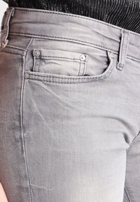 Šedé džínové kalhoty s slim fit, standardními kapsami, kovovými nýty a jemnou texturou. Pas obsahuje poutka na opasek a detaily šití.